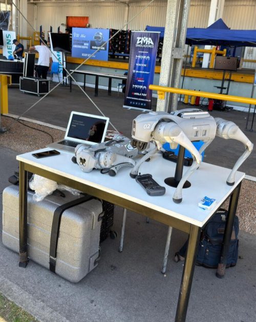 Perros robots de cuatro patas en mesa de demostración industrial con laptop y controles, listos para pruebas logísticas.